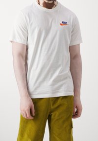 Osoba oblečená v jednoduchém bílém tričku Nike s oranžovým logem a zelených kordových kalhotách, stojící proti bílé pozadí.