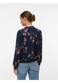 Femme aux cheveux châtains attachés en chignon, portant des boucles d'oreilles, un haut à manches longues bleu marine à motifs floraux, et un jean bleu clair, de dos sur un fond blanc.