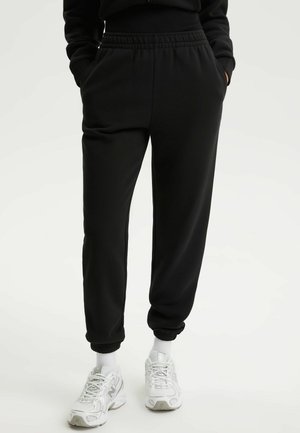 Osoba ubrana w czarne spodnie dresowe z elastycznym paskiem i ściągaczami, białe skarpetki oraz białe sportowe buty, trzymająca ręce w kieszeniach.