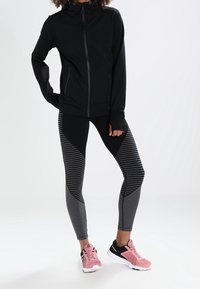 Czarna kurtka zapinana na zamek z kapturem, zestawiona z legginsami w czarno-szare paski. Obuwie z różowymi akcentami i białymi podeszwami. Gładkie tekstury na całej powierzchni.