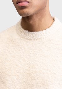 Cremfarbener Pullover mit einer weichen, flauschigen Oberfläche. Mit einem gerippten Rundhalsausschnitt. Nahaufnahme, die das Material und das Strickmuster hervorhebt.