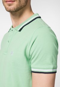Zielona koszulka polo wykonana z bawełny, z kontrastującym granatowo-białym paskiem przy kołnierzyku oraz krótkimi rękawami, z małym logo na klatce piersiowej.