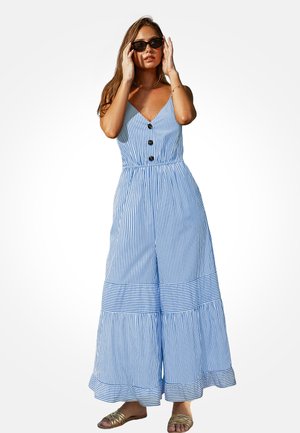 Kobieta w niebiesko-białym, pionowo paskowanym, bezrękawnym kombinezonie, poprawiająca czarne okulary przeciwsłoneczne, stojąca na tle białego tła.