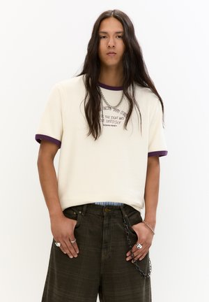 Beige kortærmet t-shirt med lilla ribbet halsudskæring og manchetter, der har tekstgrafik. Bæres med mørkegrønne bukser, der viser kæder og ringe.