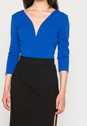 Frau trägt ein blaues Top mit V-Ausschnitt und langen Ärmeln sowie einen hoch taillierten schwarzen Rock mit seitlichem Schlitz vor einfarbigem Hintergrund.