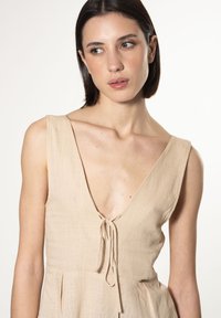 Abito in lino beige con profondo scollo a V e chiusura a laccio sul davanti. Presenta pieghe sottili e un dettaglio dorato in vita.