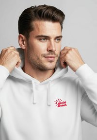 Weißer Hoodie mit einer Fronttasche und verstellbarem Kordelzug für die Kapuze. Mit rotem "Social Beach"-Logo und Sonnenstrahlen, hergestellt aus weichem Stoff.