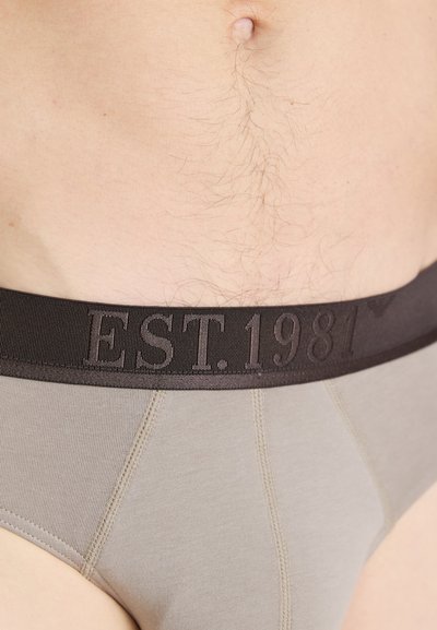 Beige herre trunks med en bred sort elastik i taljen, der har præget tekst "EST. 1981." Stoffet er glat med en tætsiddende pasform.