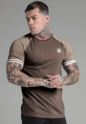 T-shirt marrone a maniche corte con maniche raglan color marrone chiaro, strisce bianche sui polsini e un piccolo logo sul petto. Texture liscia.