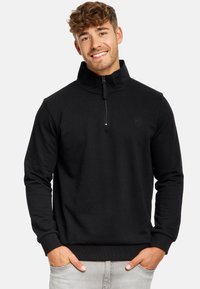 Sweatshirt zippé noir avec col haut, tissu texturé, poignets et ourlet côtelés, arborant un logo discret sur la poitrine, porté sur un jean gris.