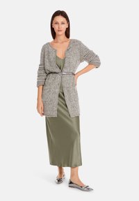 Grau gestrickte Strickjacke mit strukturiertem Finish, tailliert mit einem Gürtel, getragen über einem olivgrünen Satin-Kleid, kombiniert mit silbernen Ballerinas.