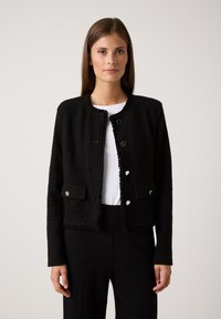 Zwarte textuurjack met een ronde hals, rafelige randen, twee voorzakken en zilveren knoopsluitingen. Draagt over een wit topje en zwarte broek.