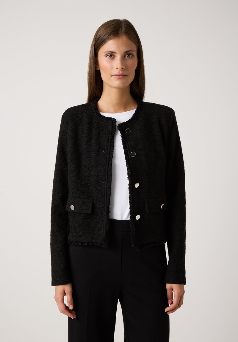 Zwarte textuurjack met een ronde hals, rafelige randen, twee voorzakken en zilveren knoopsluitingen. Draagt over een wit topje en zwarte broek.