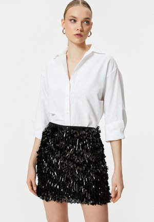 Camicia bianca con maniche arrotolate sopra una gonna nera frangiata realizzata in materiale riflettente, con accenti triangolari sovrapposti.