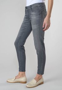 Osoba v těsných šedých džínech a béžových mokasínech, stojící proti jednoduchému světlému pozadí, ruka uvolněně na boku.