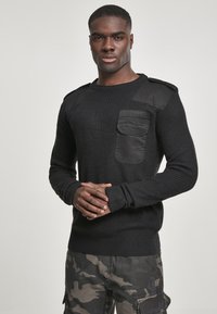 Suéter negro de punto con una superficie texturizada, que presenta un detalle en el hombro y un bolsillo en negro mate. Lleva puestos unos pantalones cargo de camuflaje.