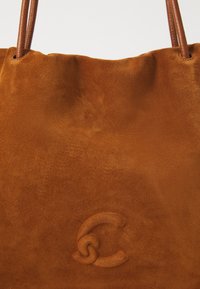 Brun semsket toteveske med preget sammenflettet "C"-logo og tynne lærstropper på en hvit bakgrunn.