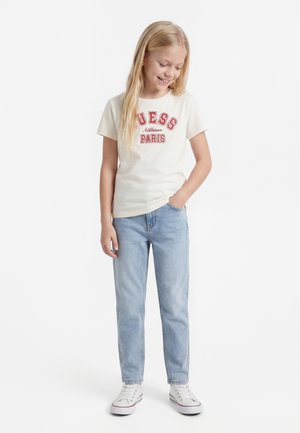 Lyshåret pige smilende, iført hvid "Guess Paris" T-shirt, lyseblå jeans, hvide sneakers, med den ene hånd i lommen, stående på hvid baggrund.