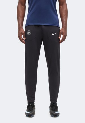 Άνδρας που φοράει μαύρη προπονητική φόρμα Chelsea FC Nike και μαύρα ποδοσφαιρικά παπούτσια Nike, στέκεται με τα χέρια χαλαρά δίπλα στο σώμα.