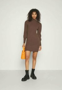 Vestido de punto marrón con cuello alto, silueta ajustada, mangas largas y un bolso naranja. Lleva botas de combate negras.