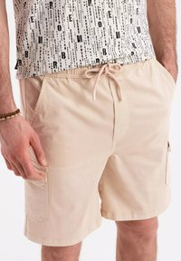 Shorts beiges avec une taille élastique et un cordon de serrage, dotés de poches latérales et d'une texture lisse. Portés avec une chemise noire et blanche à motifs.