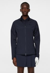 Marinblå lätt jacka med dragkedja fram, ståkrage och elastiska ärmslut. Har en diskret logotyp på bröstet.