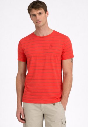 Mann trägt ein rotes kurzärmeliges T-Shirt mit dünnen horizontalen Streifen und beige Cargoshorts, steht mit einer Hand in der Tasche.