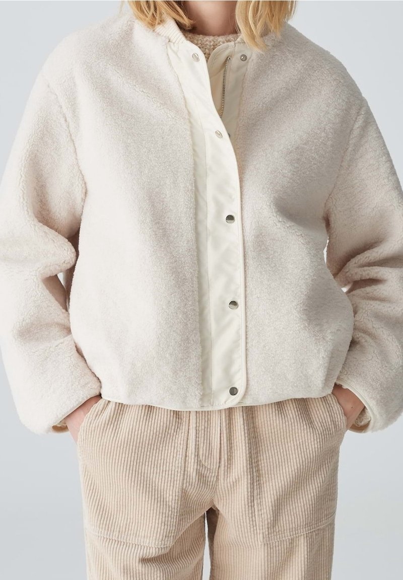 Jasno beżowa polarowa kurtka z białą plisą na napy i prążkowanym kołnierzem. Charakteryzuje się luźnym krojem i teksturowanym materiałem, w zestawieniu z sztruksowymi spodniami.
