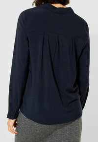 Blusa a maniche lunghe blu navy con un delicato motivo a quadri, dettaglio della piega sul retro e colletto. Texture morbida con polsini con bottoni.
