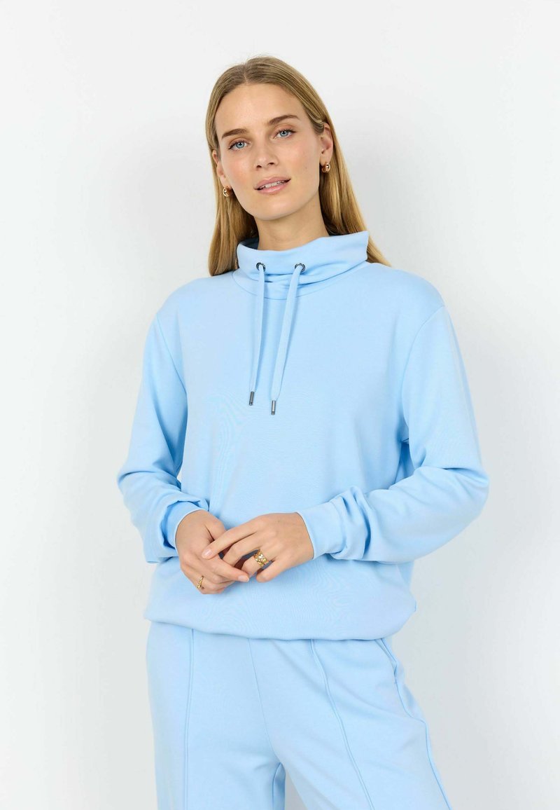 Hellblauer Pullover mit hohem Kragen, Kordelzügen und Ärmelbündchen, gefertigt aus einem weichen, strukturierten Stoff. Kombiniert mit passenden Hosen.