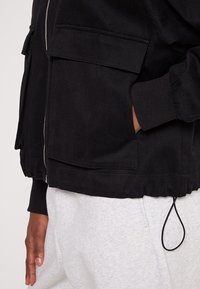 Czarna kurtka z zamkiem błyskawicznym, dwoma przednimi kieszeniami, elastycznymi mankietami i ściągaczem u dołu, wykonana z gładkiego materiału. Noszona na szarych spodniach.