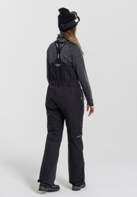 Black waterproof overall with adjustable straps, side pockets, and reinforced lower legs. Worn with a dark long-sleeve shirt and beanie.