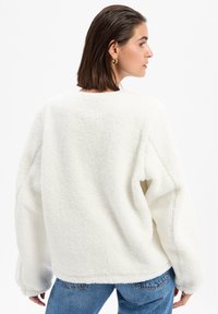 Pull en polaire blanc avec une coupe décontractée, épaules tombantes et manches longues. Texture douce, coutures minimales et col rond.