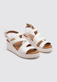 Sandalias de cuña blancas con tiras cruzadas, punta abierta y una plantilla marrón. Presenta una hebilla dorada y una suela de plataforma ligeramente texturizada.