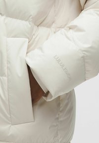 Hele kreemivärvi puffjakki, millel on tepitud disain, nähtavad õmblused, suur tasku ja särava "LULULEMON" logo varrukal.