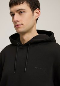 Czarna bluza z kapturem wykonana z miękkiego materiału, posiadająca przednią kieszeń kangurką, sznurki do regulacji oraz subtelnie wytłoczone logo. Gładka faktura i casualowy design.