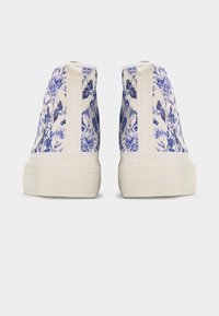 Οπίσθια όψη λευκών ψηλών αθλητικών παπουτσιών με μπλε φλοράλ και πεταλούδα σχέδιο και χοντρές εκρού λαστιχένιες σόλες σε γκρι φόντο.
