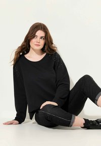 Sudadera negra con hombros decorados, cuello redondo y mangas largas, combinada con jeans oscuros que tienen un dobladillo enrollado y costuras claras.