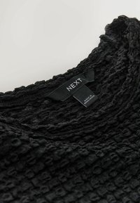 Top de punto negro texturizado con escote redondo y una etiqueta que dice "NEXT". La tela tiene un patrón en relieve y un acabado suave.