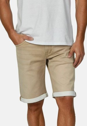Uomo che indossa pantaloncini beige risvoltati e una maglietta bianca, mano destra in tasca, in piedi davanti a uno sfondo uniforme.
