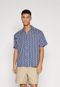 Camisa de manga corta en tejido azul con un patrón geométrico blanco, diseño con cuello y un pequeño logo verde en el pecho.