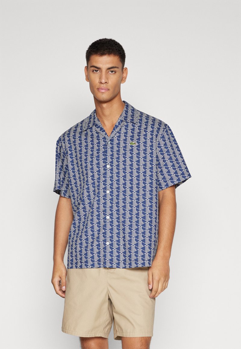 Camisa de manga corta en tejido azul con un patrón geométrico blanco, diseño con cuello y un pequeño logo verde en el pecho.