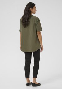 Camisa de manga corta verde oliva con dobladillo redondeado, combinada con pantalones negros ajustados y bailarinas negras. Textura de tela suave, diseño casual.
