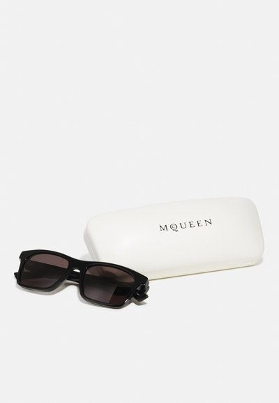 Occhiali da sole neri rettangolari con lenti oscurate accanto a un astuccio bianco rettangolare con "MQUEEN" in testo nero.