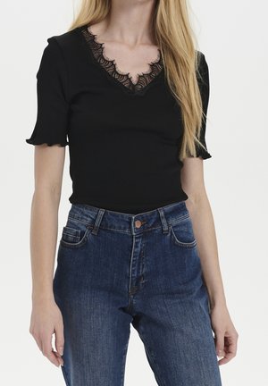 Kvinde iført en sort kortærmet top med V-hals med blondekant og højttaljede blå denimjeans mod en ensfarvet baggrund.