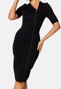 Frau in einem figurbetonten schwarzen Midi-Kleid mit kurzen Ärmeln und einem durchgehenden Reißverschluss auf der Vorderseite, posierend mit der Hand auf der Hüfte.