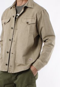 Giacca di cotone beige con chiusura a bottoni, due tasche al petto e una patch del logo. Maniche lunghe con polsini, indossata sopra una camicia grigia.