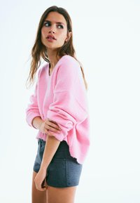 Różowy, oversize'owy sweter z dekoltem w serek i prążkowanymi mankietami, zestawiony z ciemnoszarymi szortami z guzikami. Miękka faktura, swobodny krój.