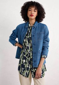 Veste en denim bleu moyen avec fermeture à boutons et poche avant, associée à une chemise à motif avec des fleurs navy sur un fond blanc et vert.