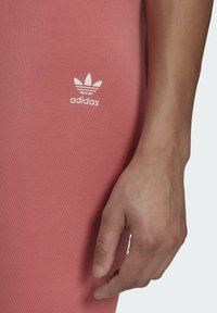 Růžové sportovní legíny z pružného materiálu, na kterých je v blízkosti boků bílá značka Adidas, s detailním pohledem na ruku položenou na tkanině.